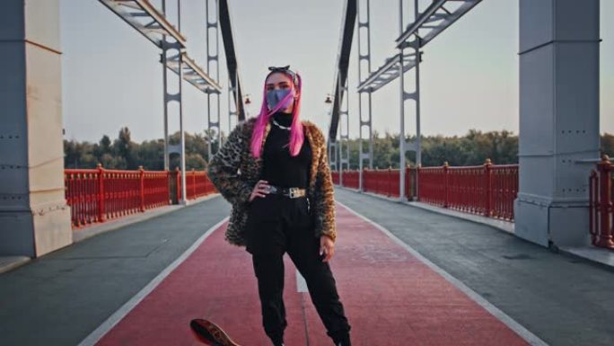 穿着非正式服装和防护面具的粉红色头发的时髦女士正站在滑板桥上摆姿势