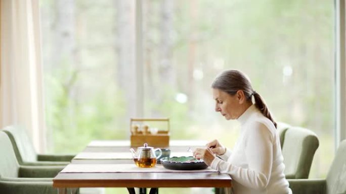 侧视图缩小了白发的高级女性的镜头，她独自坐在咖啡馆的桌子上，吃饭，边吃饭边思考