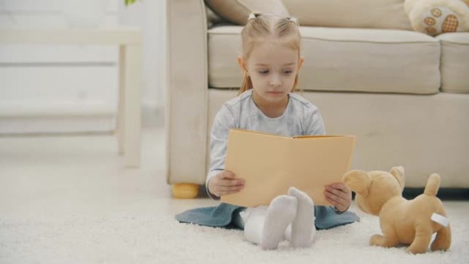 4k视频的金发小女孩手里拿着一本书坐在软玩具旁边。