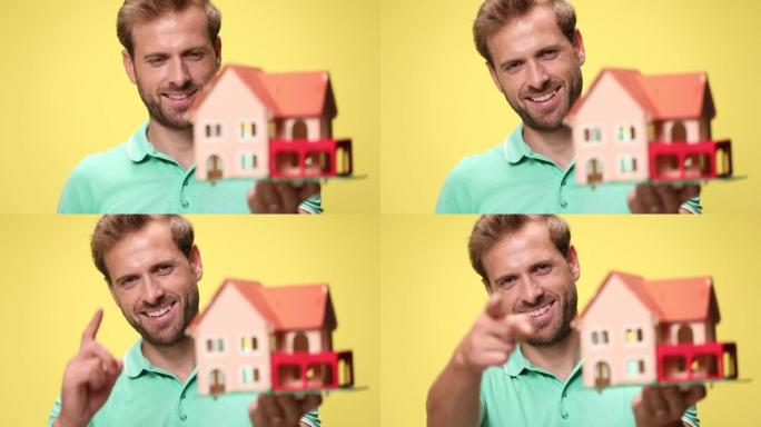 微笑的年轻休闲小伙子举起房子模型，在黄色背景上推荐和指指点点