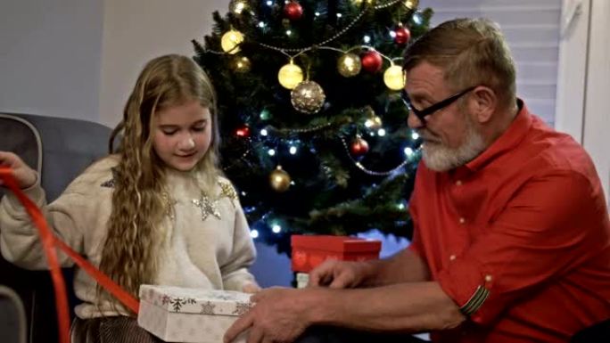 圣诞树附近的祖父和孙女。女孩打开礼物，欢欣鼓舞。新年快乐