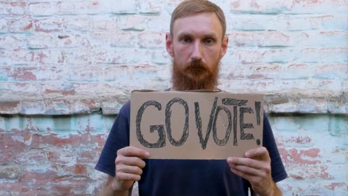 一名男子在城市背景的砖墙上展示写着“去投票”的纸板。投票的概念。做出政治选择，用你的声音。一个戴着防
