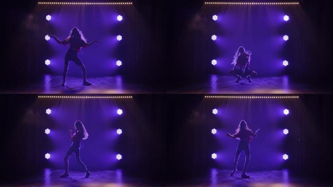 年轻的运动女孩正在积极跳舞时髦的爵士乐。在黑暗的工作室中，蓝灯背景下的剪影。慢动作