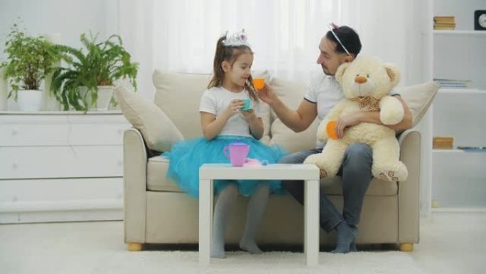 父亲、女儿和泰迪举行茶会。他们坐在沙发上，喝茶。有趣的父亲和可爱的皇冠女孩。