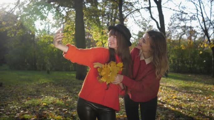 秋天的季节，美丽的微笑的年轻女性朋友在手机上自拍，在阳光下晒着一束黄色的叶子，在秋天的树叶上联合散步