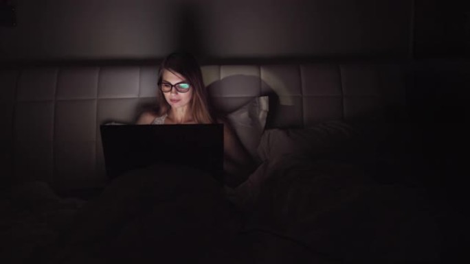 戴眼镜的女人在黑暗中在笔记本电脑上工作。一个女孩在深夜花时间在电脑上的肖像