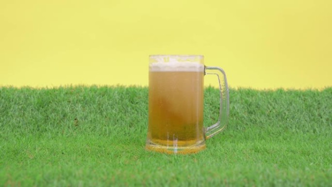 将冷清爽的泡沫啤酒倒入大杯子中，杯子站在人造草坪的绿色草地上，黄色背景，正面视图。小玩具足球滚出。
