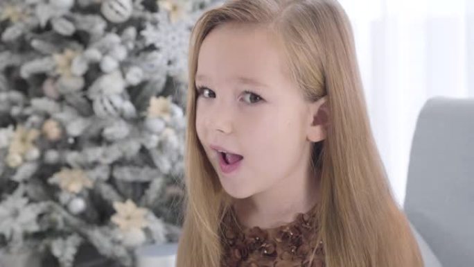 相机在可爱的长发白人女孩的脸上移动。除夕坐在圣诞树旁边的漂亮孩子。美丽，假期。