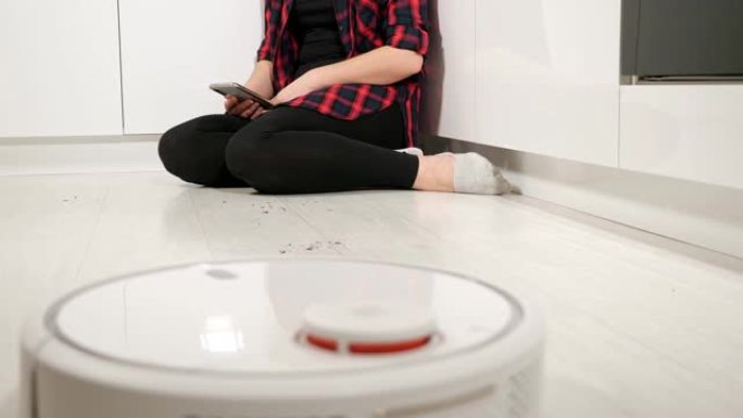 无法识别的带电话的女人坐在地板上，而机器人吸尘器清洁地板