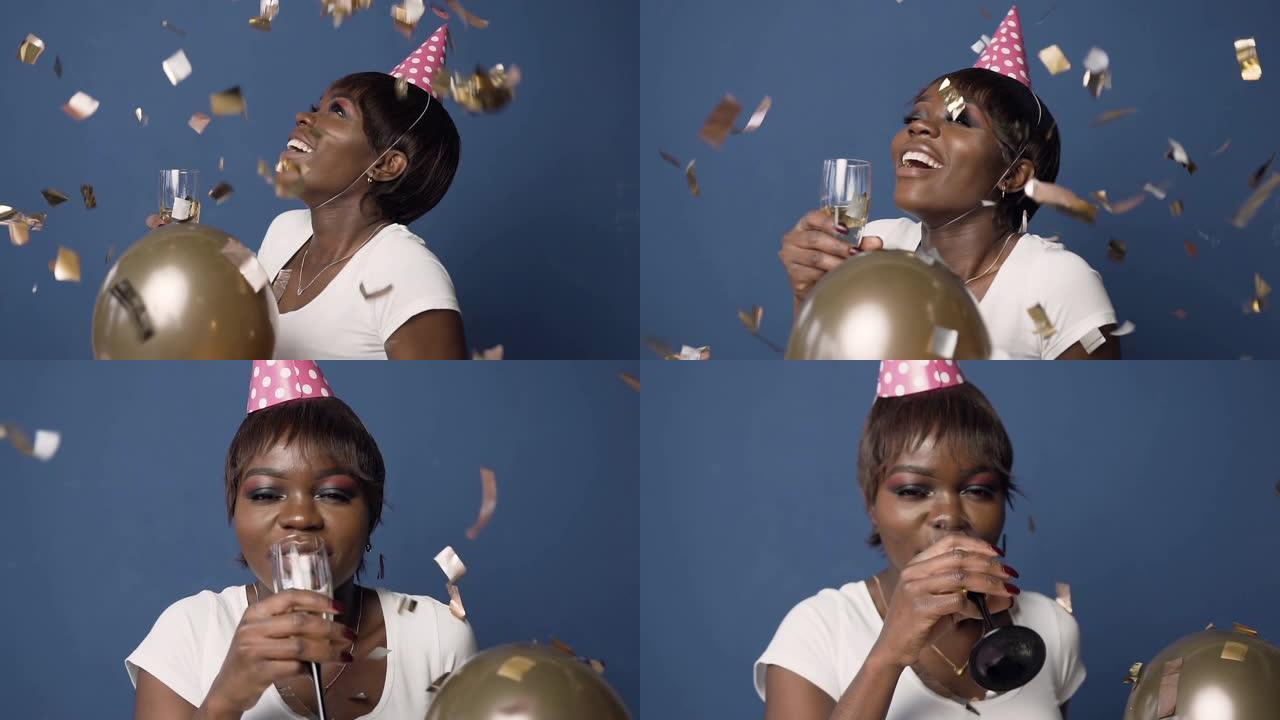 快乐的非洲女孩戴着帽子，一只手拿着香槟玻璃，另一只手拿着气球，看着蓝色背景上飞落的五彩纸屑