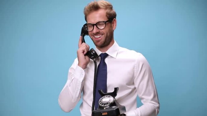 年轻的商人拿着一个复古的电话，拿起，说话，大笑，挂断并指着蓝色背景上的相机