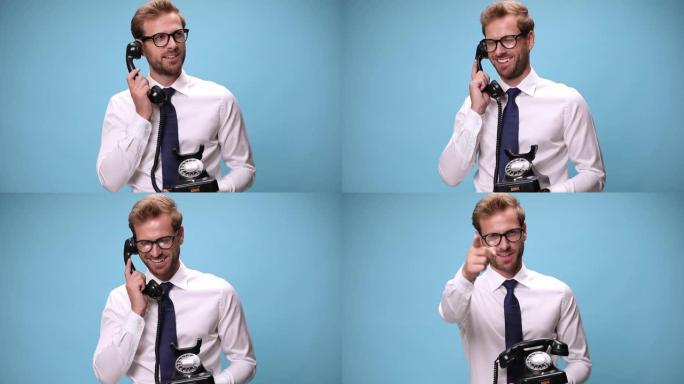 年轻的商人拿着一个复古的电话，拿起，说话，大笑，挂断并指着蓝色背景上的相机