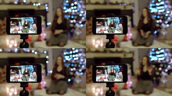 漂亮的年轻女子坐在令人惊叹的圣诞节背景上，用智能手机摄像头为在线观众录制视频。有趣的社交媒体视频信息