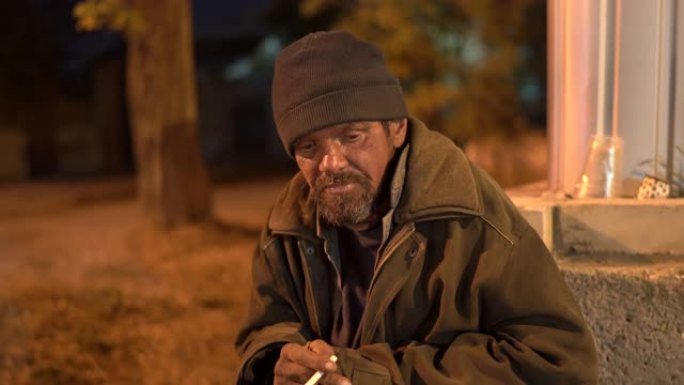 悲伤的流浪汉晚上坐在街上。孤独寒冷的乞丐吸烟