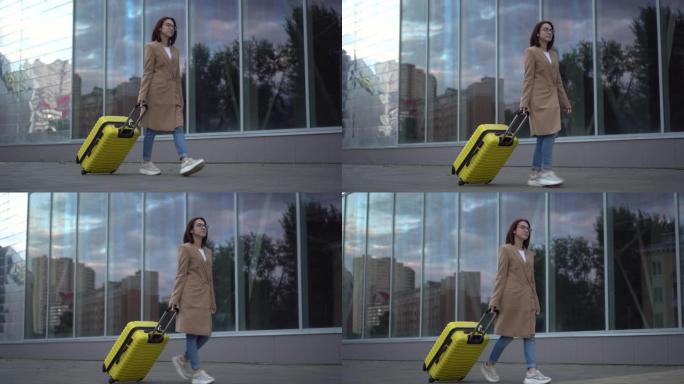一个年轻的女人带着一个黄色的手提箱走路。一个戴着眼镜和外套的女孩沿着镜子的建筑物行走。