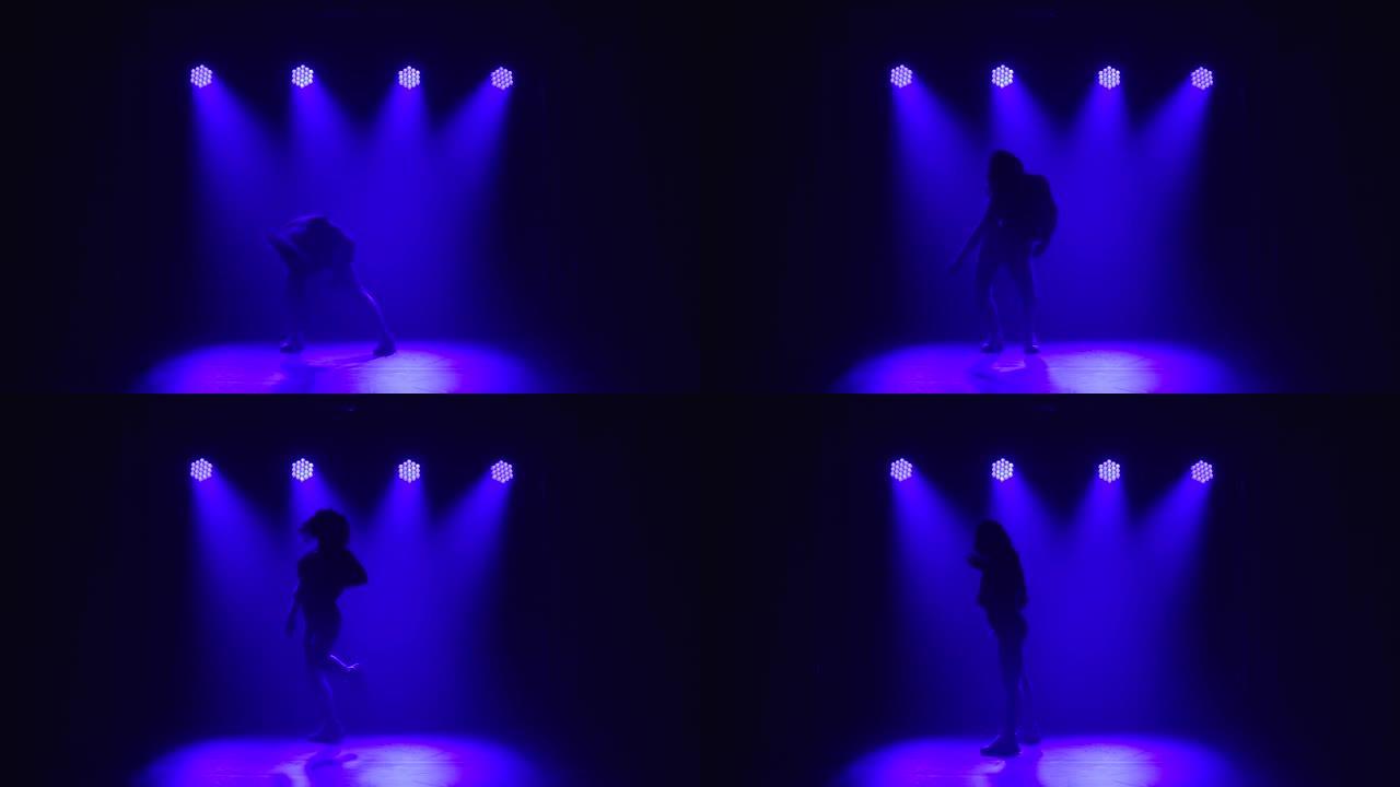 一个波浪形头发的女孩专业跳舞爵士乐放克元素。在上方蓝色灯光的背景下，在黑暗的工作室中舞者的剪影。慢动