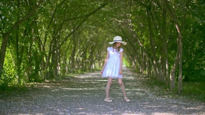 戴着帽子的无忧无虑的卷发女孩在夏天盛开的小巷背景上扭曲。快乐女孩少年微笑着，在夏日公园绿色开花的树木