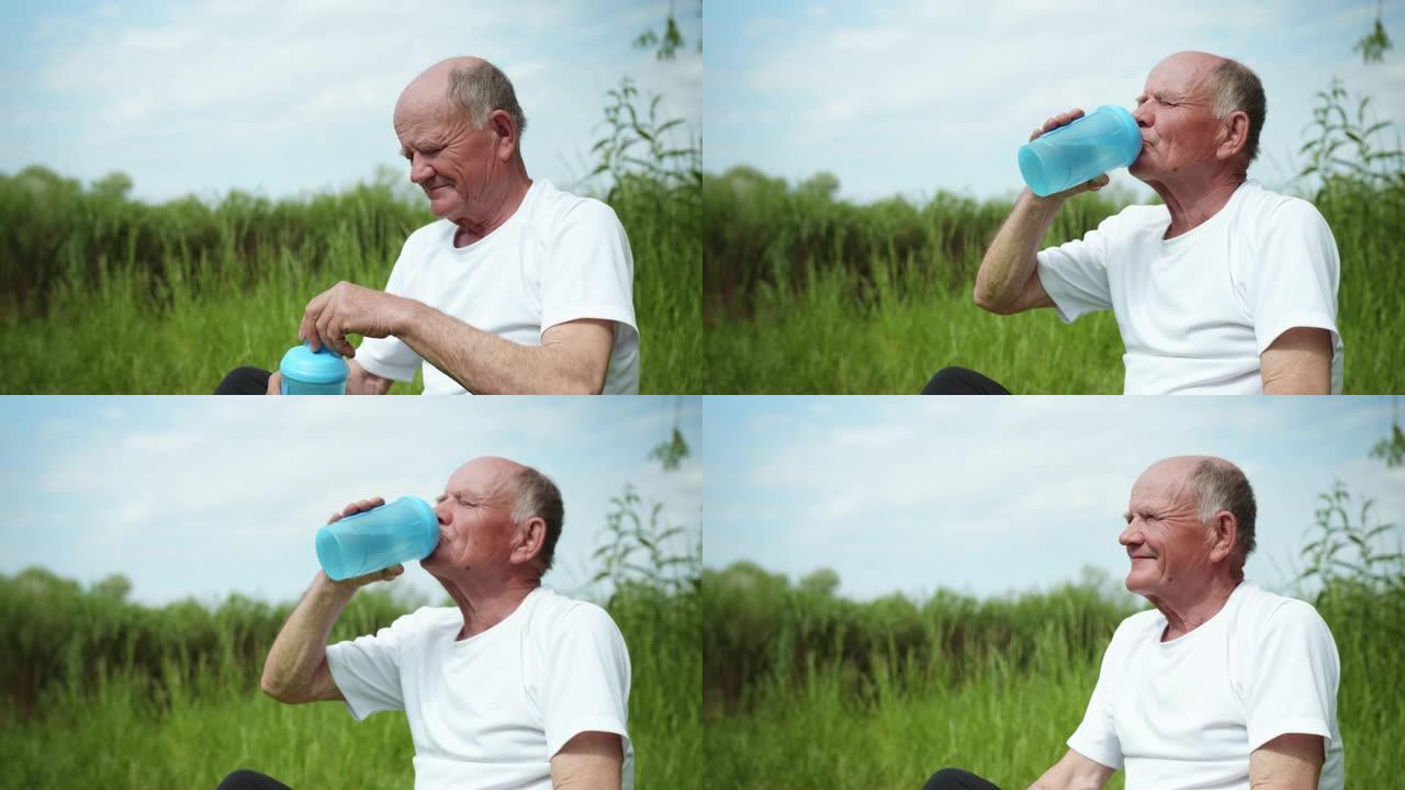 健康的老人恢复平衡，在运动或冥想后，坐在绿色植物的露天背景下的垫子上，从瓶子里喝干净的水，用于运动营
