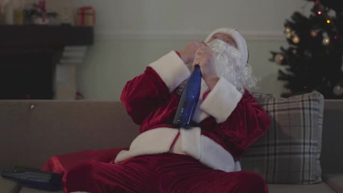 穿着圣诞老人服装的醉酒老人坐在沙发上喝酒。孤独的高加索人独自度假。坏圣诞老人，酗酒，消极主义。