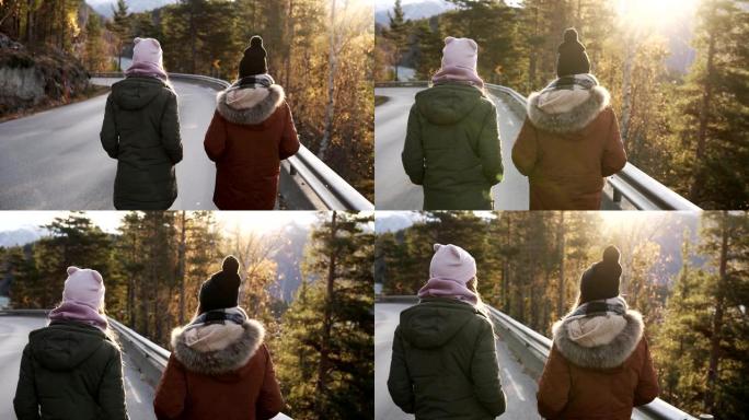 两名女游客在一条穿过乡村的漫长道路上慢动作行走，走向背景上的雪山山峰和金色树木。罕见的观点