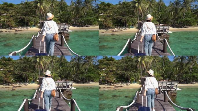 当她乘船到达泰国的特拉特时，带着行李的美丽的亚洲女人走在夏日海滩酒店区前的木制码头上。旅游、暑假和度