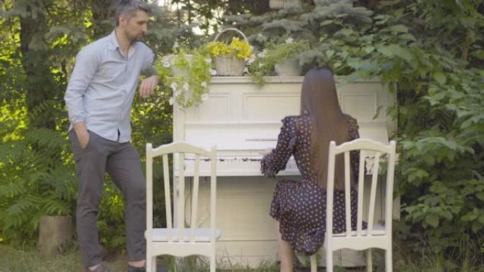 快乐的成年男子站在钢琴旁边，听着黑发女人在演奏音乐。积极富有创造力的高加索夫妇在户外度过周末的肖像