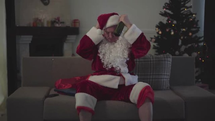 打扮成圣诞老人的白人男子戴上红帽子并打开电视。疲惫的老家伙端着一瓶啤酒坐在沙发上。
