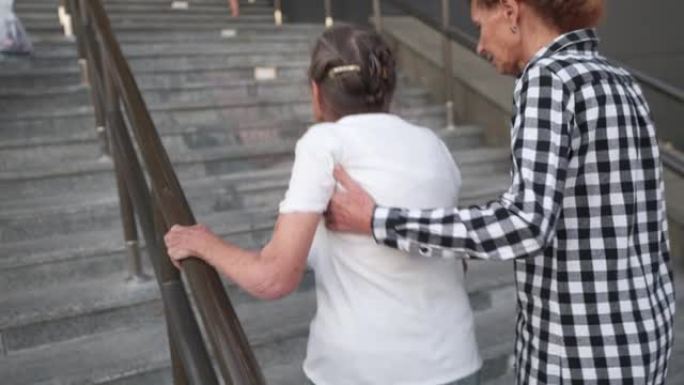 高级妇女和她的中年女儿走在高层建筑的院子里。护士帮助一个老养老金领取者在外面的楼梯上爬。照顾者帮助、