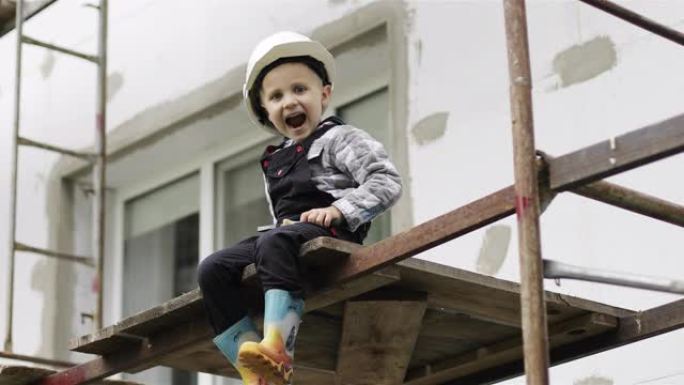 一个戴着白色头盔的小孩建筑商坐在脚手架上，手里拿着锤子和钳子。