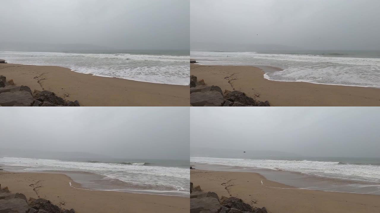 在恶劣的天气，下雨天，深灰色和刮风的日子里，海浪击中了海滩的沙滩，而鸟儿飞过的背景冲浪者可以看到试图