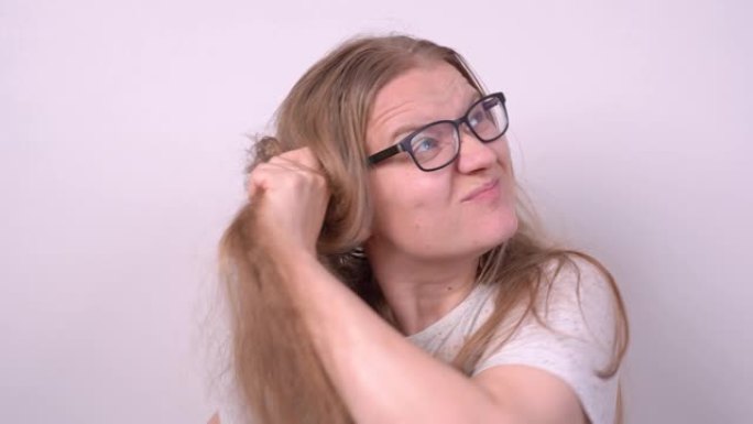 戴眼镜的可爱金发女孩试图用木刷梳理稀薄纠结的长发，在白色背景上做一个时尚的发型，但没有成功。护理问题