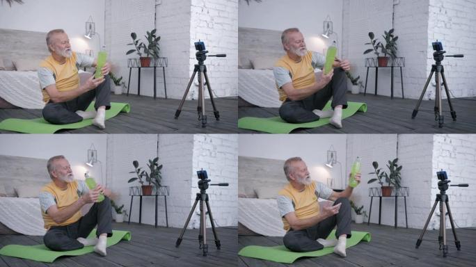 老博主，衰老的影响者喜欢运动在智能手机上进行饮料评论vlog向订阅者推荐坐在创意室内瑜伽垫上的水