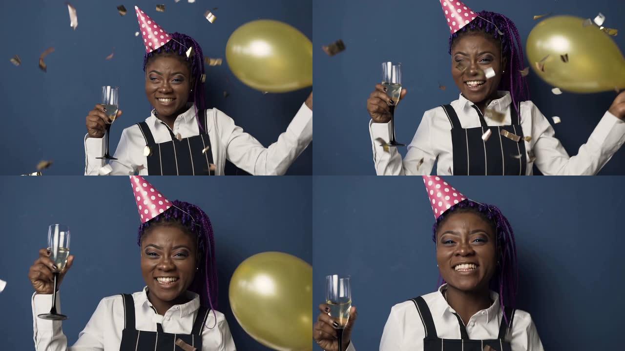 快乐的年轻非洲女人戴着生日帽，一只手拿着香槟玻璃，另一只手拿着气球跳舞，五彩纸屑在蓝色背景上飞来飞去
