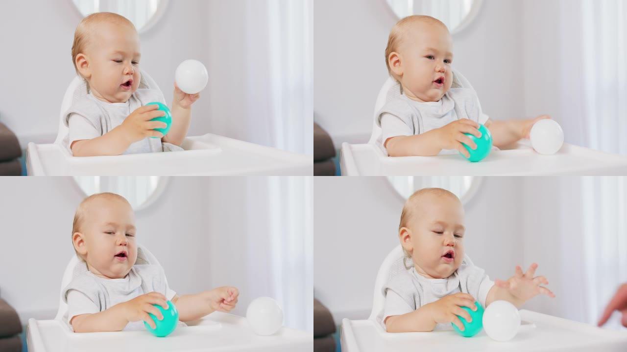白色高高的婴儿椅子上的小婴儿特写镜头，他双手握着两个塑料球-绿色和白色-并将它们相互撞击。当失去一个