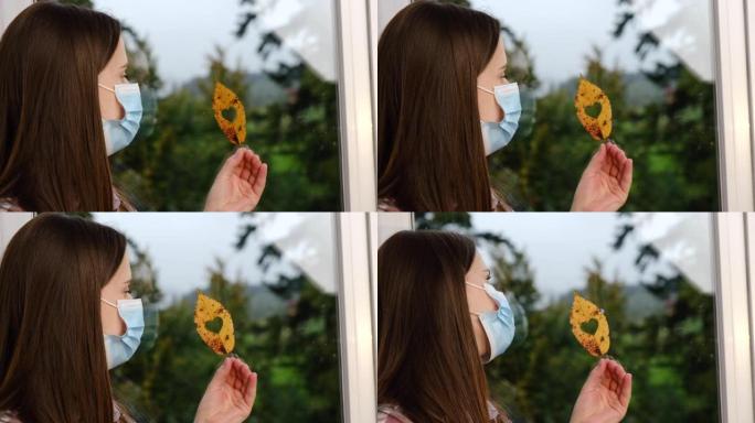 坐在窗台上戴着医用蓝色口罩的年轻女子的特写肖像，上面放着带有心形孔的黄色小叶，在检疫时在家中发现。冠