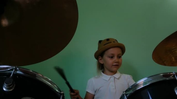 在音乐学校学习鼓的孩子。黑色鼓包。绿色墙壁背景上的鼓刷训练小音乐家鼓手。戴着帽子的金发小女孩在课上玩