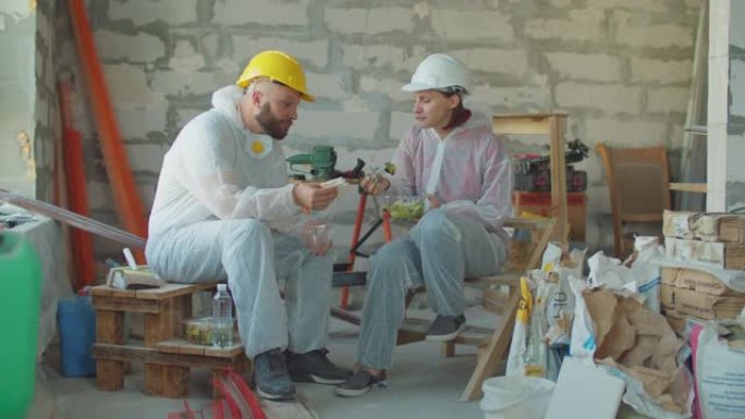 两名画家工人在建筑工地吃午饭。戴着安全帽的男人和女人坐在建筑材料旁，边吃饭边聊天。