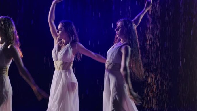 三个穿着白色长裙的女人在冒烟的背景下在雨中与玫瑰共舞。慢动作。特写