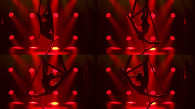 一位专业的空中飞人借助空中丝绸在空中旋转。红色霓虹灯中苗条的年轻女子的轮廓。特写