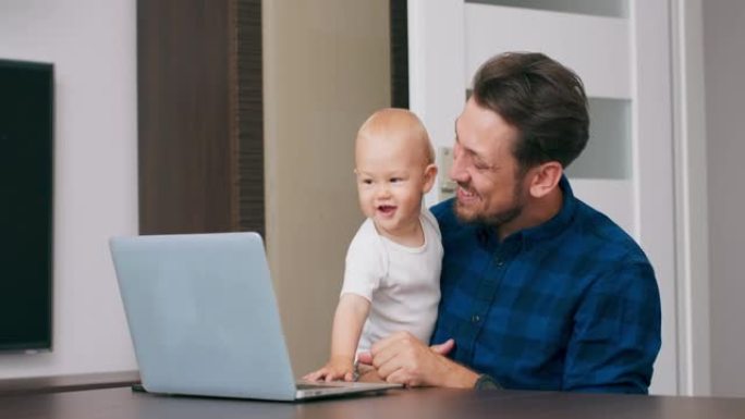 年轻的大胡子男人坐在家里的桌子上，拿着笔记本电脑，膝盖抱着可爱的婴儿，通过通讯器与婴儿的母亲交谈。男