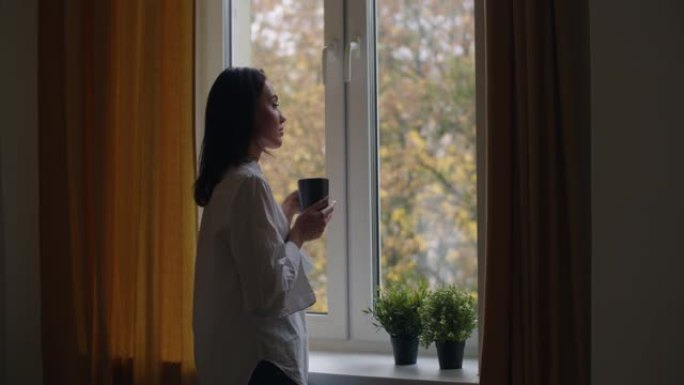 沉思女孩的侧面肖像站在窗边，喝杯茶慢动作。年轻女子喝热咖啡，看着外面的秋天风景复制空间。孤独抑郁孤独