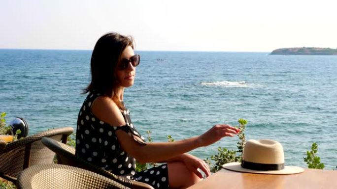 黑发女孩坐在海边咖啡馆的桌子旁，欣赏海景。炎热的夏日