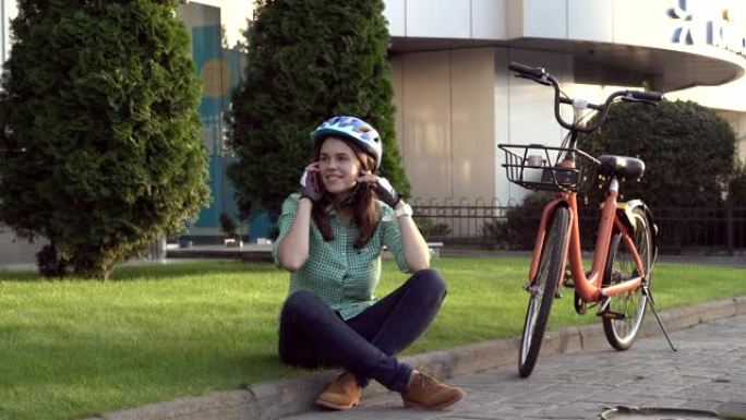 人和城市滚动自行车，环保运输。坐在草地上休息的年轻白人女工在橙色自行车附近使用红色手机