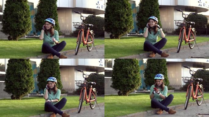 人和城市滚动自行车，环保运输。坐在草地上休息的年轻白人女工在橙色自行车附近使用红色手机