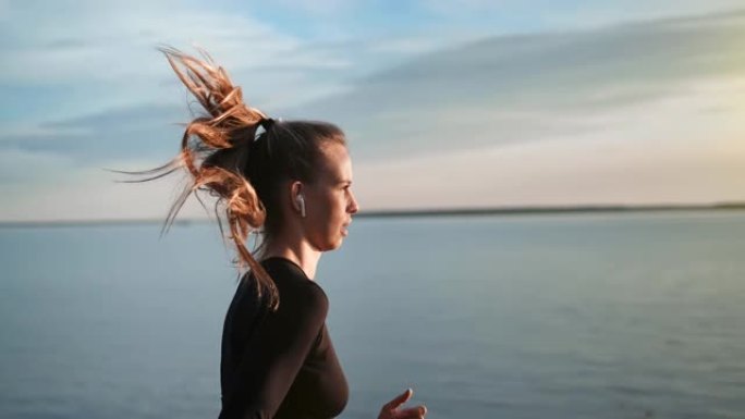 运动女运动员戴着耳机在海滩上跑步在日落慢动作。4k Dragon RED摄像机