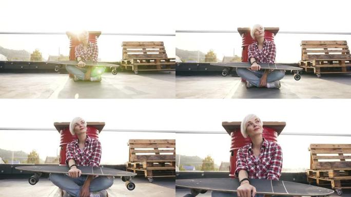穿着牛仔裤和红色方格衬衫的年轻时髦女孩坐在现代工业建筑屋顶的地板上，拿着滑板。夏日，金发女孩坐在户外