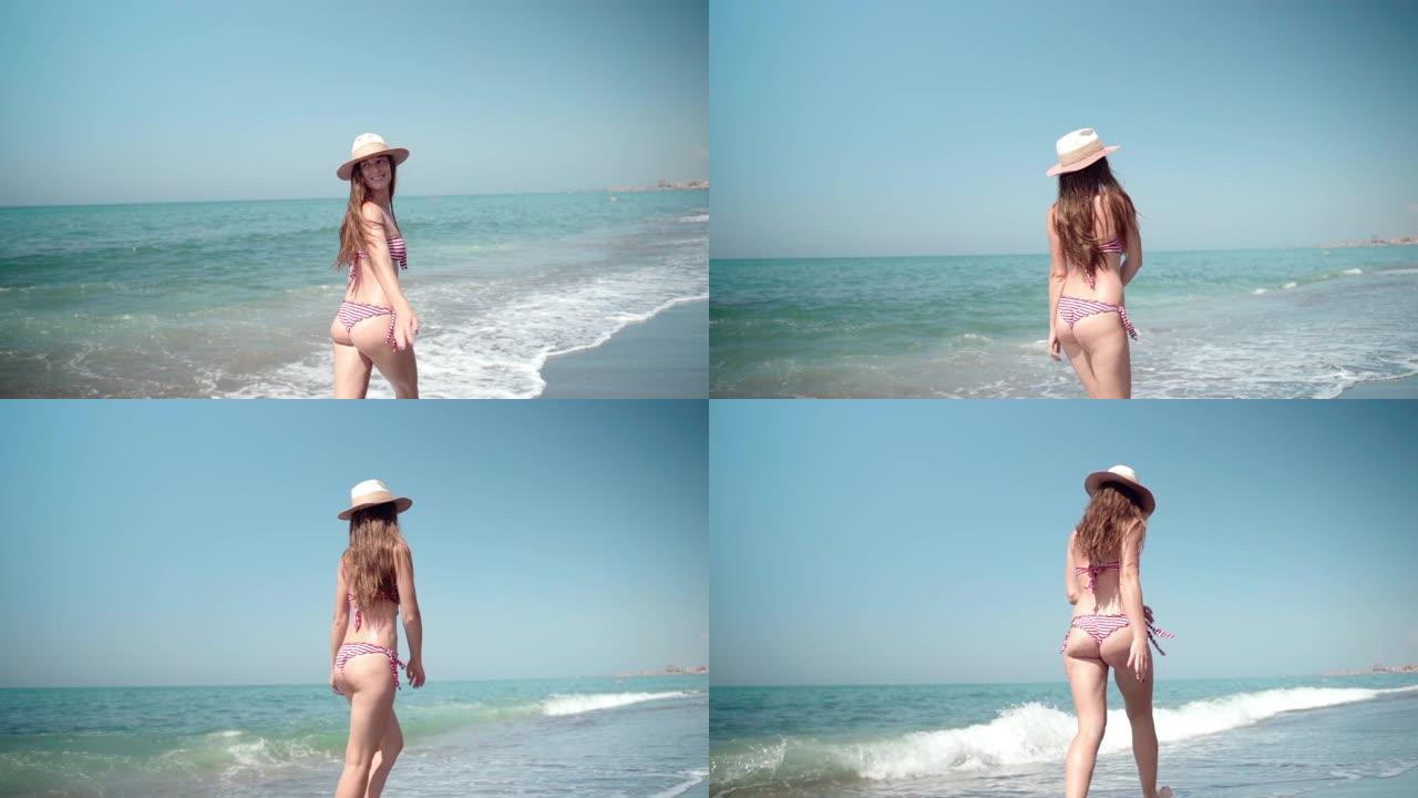 穿着夏日帽子和诱人比基尼的迷人性感黑发女孩沿着海边奔跑，转身微笑。身材迷人的漂亮女孩享受假期和早晨漫