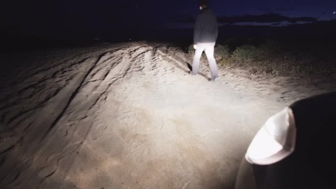 一个人在晚上走在沙滩上，远离汽车的前灯