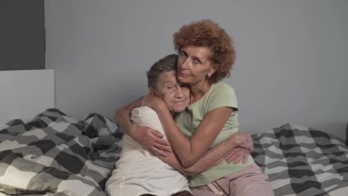 成年孙女抚摸她的额头，拥抱一位年迈的祖母，在卧室里享受着温柔的时刻。可爱的女儿在公寓里用爱拥抱她快乐