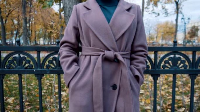 一个穿着淡紫色外套，系着皮带的女孩站在公园的篱笆上。外套特写。慢动作。